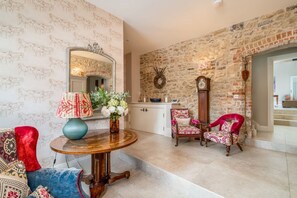 A room with a stone wall, a round wooden table with a lamp, a mirror, a red chair, and a clock. - The Stables, Oxfordshire 4 bed (Oxford)
