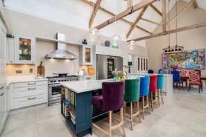 A modern kitchen with a central island, wooden beams, and colorful bar stools. - The Stables, Oxfordshire 4 bed (Oxford)