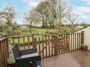 Eine Holzterrasse mit einem Korbstuhl und einem Weinglas, mit Blick auf einen Garten mit Bäumen und einem Gewächshaus.
