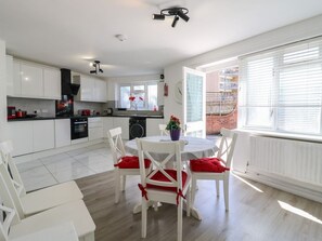 A modern kitchen with white cabinetry, a dining area with white chairs and red cushions, and a large window with blinds. - The Alex: Modern Living in Deptford (Deptford)