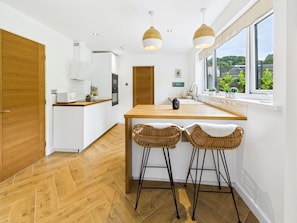 A modern kitchen with a wooden island, two wicker bar stools, and a view through a large window.