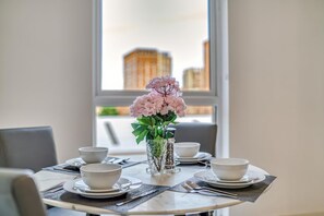 Ein Esstisch ist gedeckt mit weißen Schalen und Tellern, eine klare Vase mit rosa Blumen und ein Fenster mit Blick auf die Stadt.