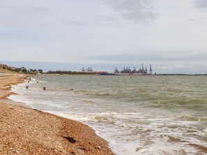 Ein Strand mit Kieselsteinen, ruhigem Wasser und im Hintergrund Industriegebäuden.