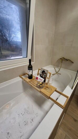 A bathroom with a bathtub, a window, a wooden tray with a bottle, chocolates, and a wine glass. - Holiday House Likarija ⭐️⭐️⭐️⭐️ (Lički Novi)