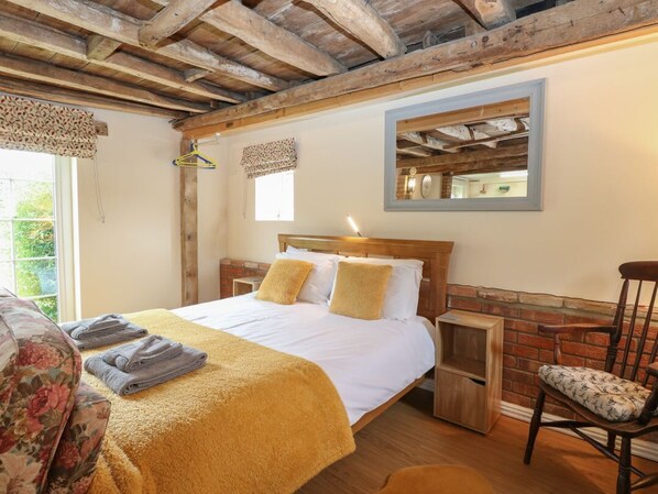 A bedroom with a wooden bed, a nightstand, a chair, and a window with a view of greenery. - Pear Tree Barn, Stalham (Norwich)
