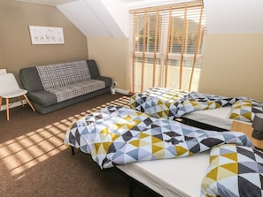 A room with a sofa, two beds with patterned bedding, a small table, and a window with blinds.