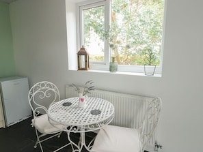 A small dining area with a round table and white chairs, a window with a view of trees, a radiator, and a small cabinet.