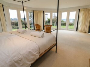 Ein Schlafzimmer mit einem Himmelbett, zwei Sesseln und großen Fenstern mit Vorhängen.