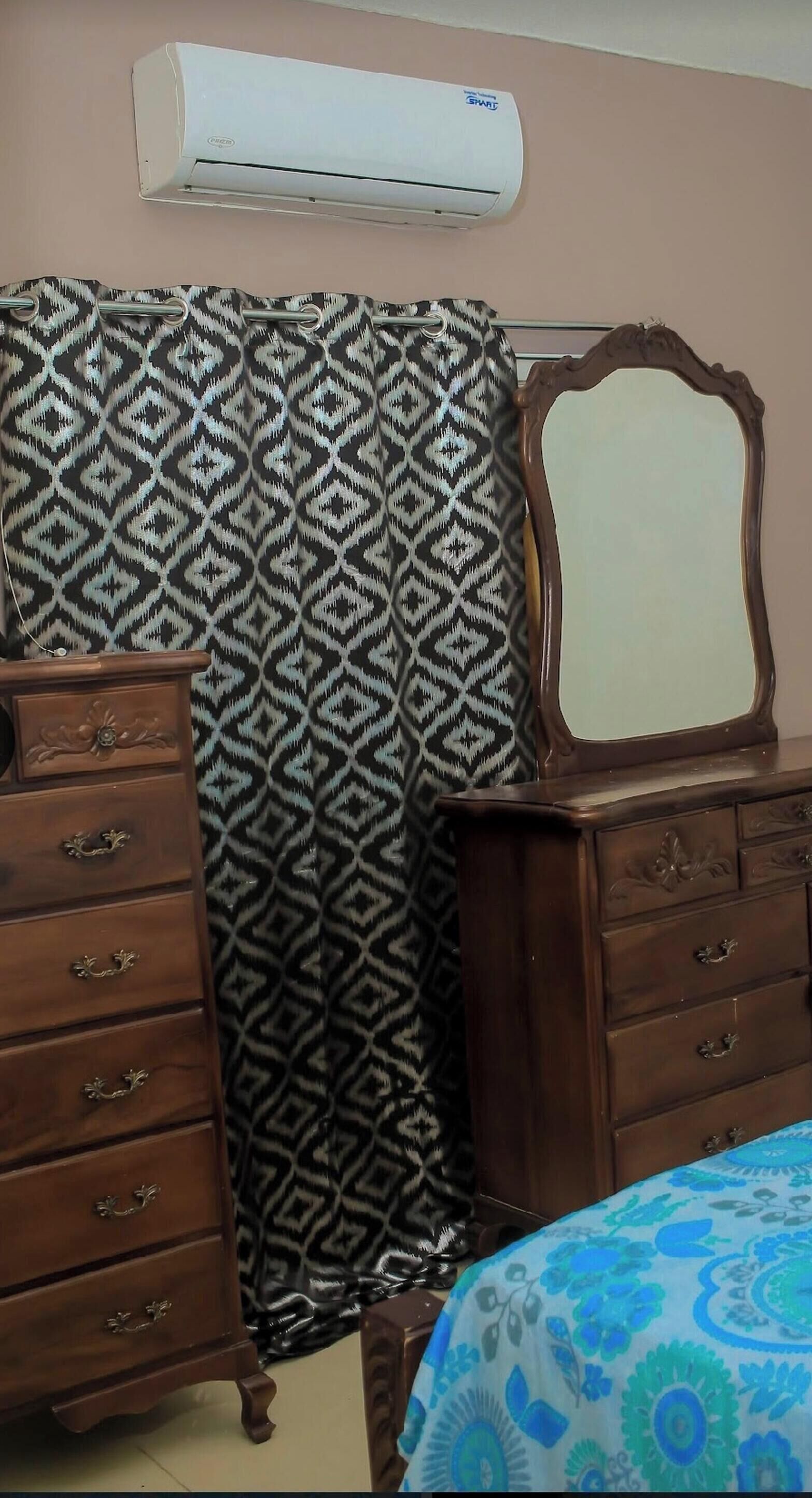 A bedroom with a patterned curtain, a wooden dresser, a mirror, and an air conditioning unit.