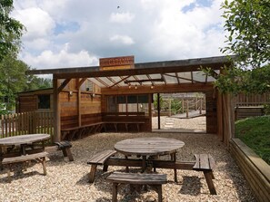 Et udendørs siddeområde med træborde og bænke, en overdækket terrasse, og et skilt med teksten 'The Cabana'.