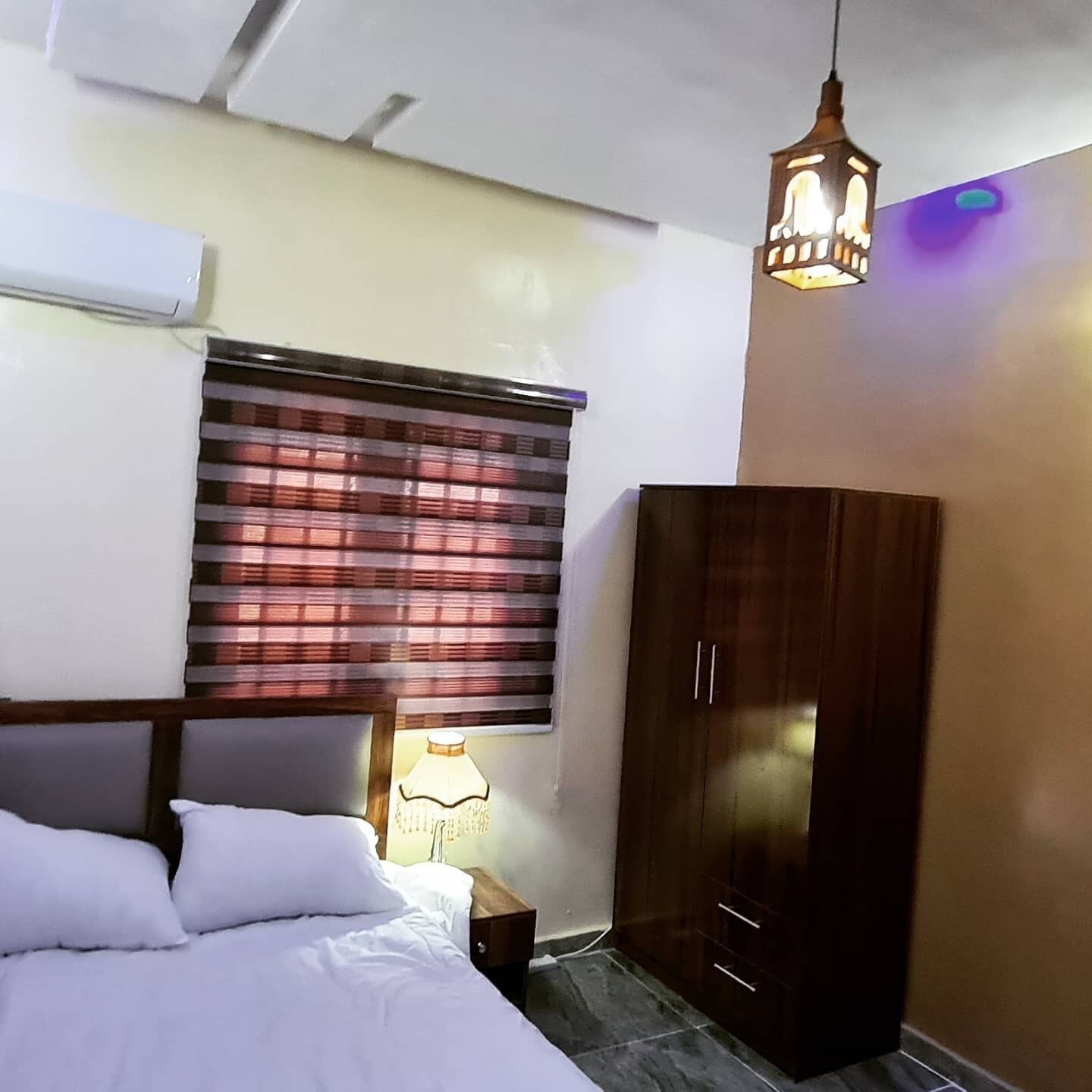 A bedroom with a bed, a wardrobe, a window with blinds, and a hanging light fixture.
