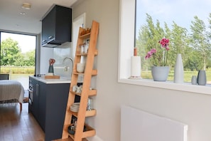 A modern kitchen with a wooden ladder shelf, a large window with a view of greenery, and a dining area with a table and chairs. - Riverside Exclusive Retreat, St Columb Major (Newquay)