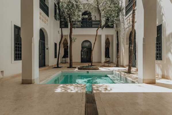 Un espace piscine offrant une vue dégagée sur le bassin, entouré de murs et de colonnes blancs, avec des arbres et un balcon en arrière-plan.