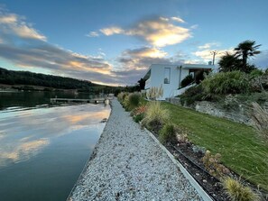 A riverside walkway with a modern building, a calm water body, and a clear sky.