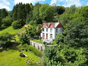 A white house with a red door, surrounded by lush greenery and a stone bridge. - The Woodlands, Berrynarbor (Ilfracombe)