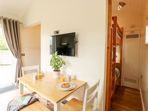 Een eetgedeelte met een houten tafel, stoelen, een potplant en een aan de muur bevestigde televisie.