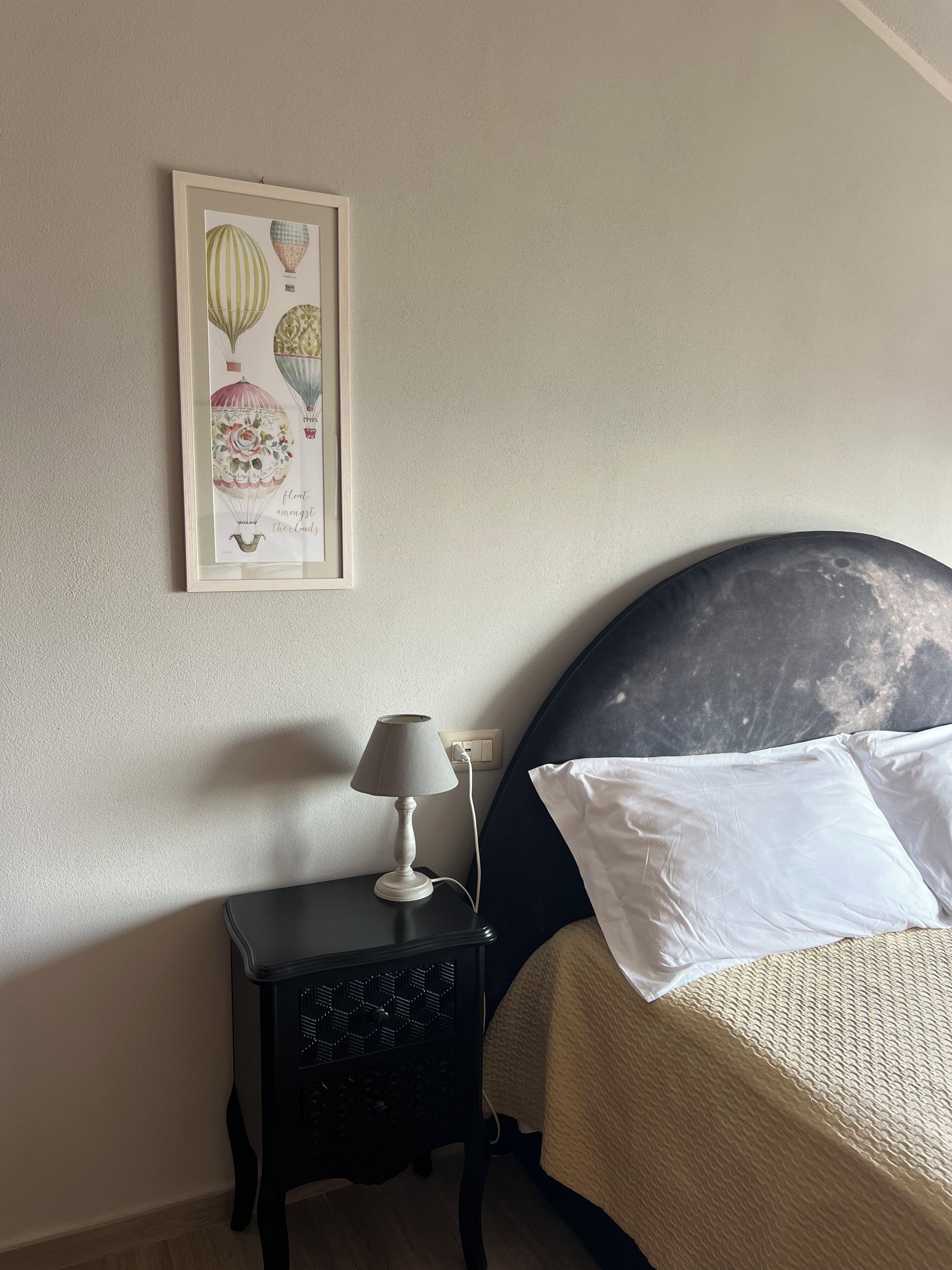 Un dormitorio con una cama, una mesita de noche con lámpara, un cuadro en la pared y un enchufe.