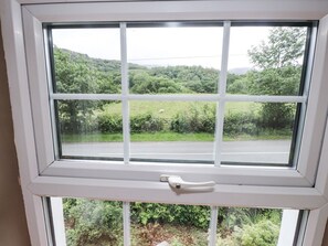 A window with a white frame and a white handle, offering a view of a green landscape with trees and a road. - Sygun Cottage, Nant Gwynant (Caernarfon)