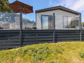 A modern building with a glass facade, a wooden deck, and a patio area with a chair and umbrella. - 63 The Maples, Sticker, 2 bed. (St Austell)