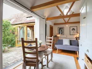 A cozy room with a wooden ceiling, a dining table with chairs, a sofa, and a window overlooking a garden. - Charming Little Barn Retreat in Orwell (Royston)