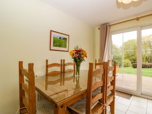 A dining area with a glass table and wooden chairs, a vase with flowers, a framed picture on the wall, and a view through a glass door. - Dovecote Cottage, Gloucester (Gloucester)