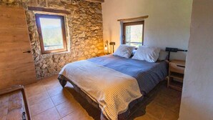 Ein Schlafzimmer mit Steinwand, Holzbalkendecke, einem Bett mit Decke, zwei Nachttischen und einem Fenster.