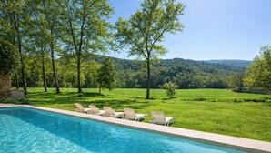 Ein Swimmingpool mit klarem, blauem Wasser, umgeben von einer Rasenfläche und Bäumen, mit Blick auf einen bewaldeten Hügel im Hintergrund.