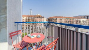 Un balcon avec une table et des chaises rouges, donnant sur un paysage urbain composé d’immeubles et d’un ciel dégagé.