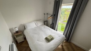 A bedroom with a bed, a nightstand, a radiator, a window with curtains, and a view of greenery outside.