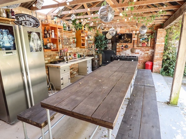 Eine rustikale Außenküche mit Holztisch, Kühlschrank und diversen Barutensilien.