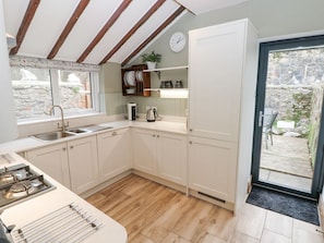 A kitchen with white cabinets, a sink, a stove, and a window overlooking a stone wall. - Aubrey Villa in Pembrokeshire. (Tenby)