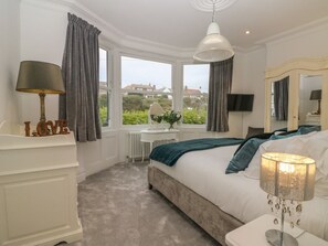 Ein Schlafzimmer mit Bett, Nachttisch, Lampe, Fenster mit Vorhängen und Blick ins Grüne.