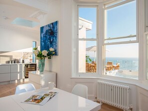 Ein modernes Zimmer mit einem großen Erkerfenster, einem Esstisch mit einer Zeitschrift, einer weißen Vase mit Blumen und Blick auf das Meer.