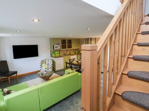A living room with a green sofa, a TV mounted on the wall, and a kitchen in the background. - Lime Cottage, Aylesbeare (Exeter)