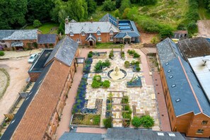 An aerial view of a courtyard with a central fountain, surrounded by brick buildings with multiple windows and a sloped roof. - Lime Cottage, Aylesbeare (Exeter)