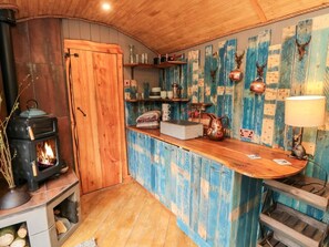 Ein rustikales Interieur mit Holzofen, Regalen mit diversen Gegenständen und einer blauen Wand mit dekorativen Elementen.