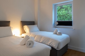 Ein Hotelzimmer mit zwei Betten, einem Fenster mit Blick auf Grünflächen und einem Nachttisch mit Lampe.