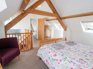 A bedroom with a wooden ceiling, a bed with a floral bedspread, a purple chair, and a view of the sea through a window. - Woodpecker Cottage, Llangollen (Llangollen)