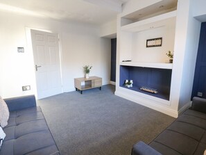 A living room with a grey carpet, a dark sofa, a small wooden cabinet, and a fireplace with a mantel. - 16 Salvin Terrace, Fishburn (Stockton-on-Tees)