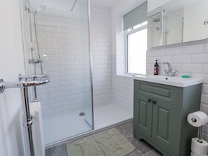 Un baño moderno con cabina de ducha de vidrio, paredes de azulejos blancos, un mueble de tocador verde con espejo y una ventana que aporta luz natural.