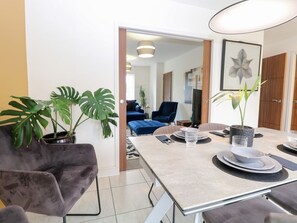 A modern dining area with a table set for two, potted plants, and a view into a living space with a blue sofa. - Manor Park View, Norwich (Norwich)