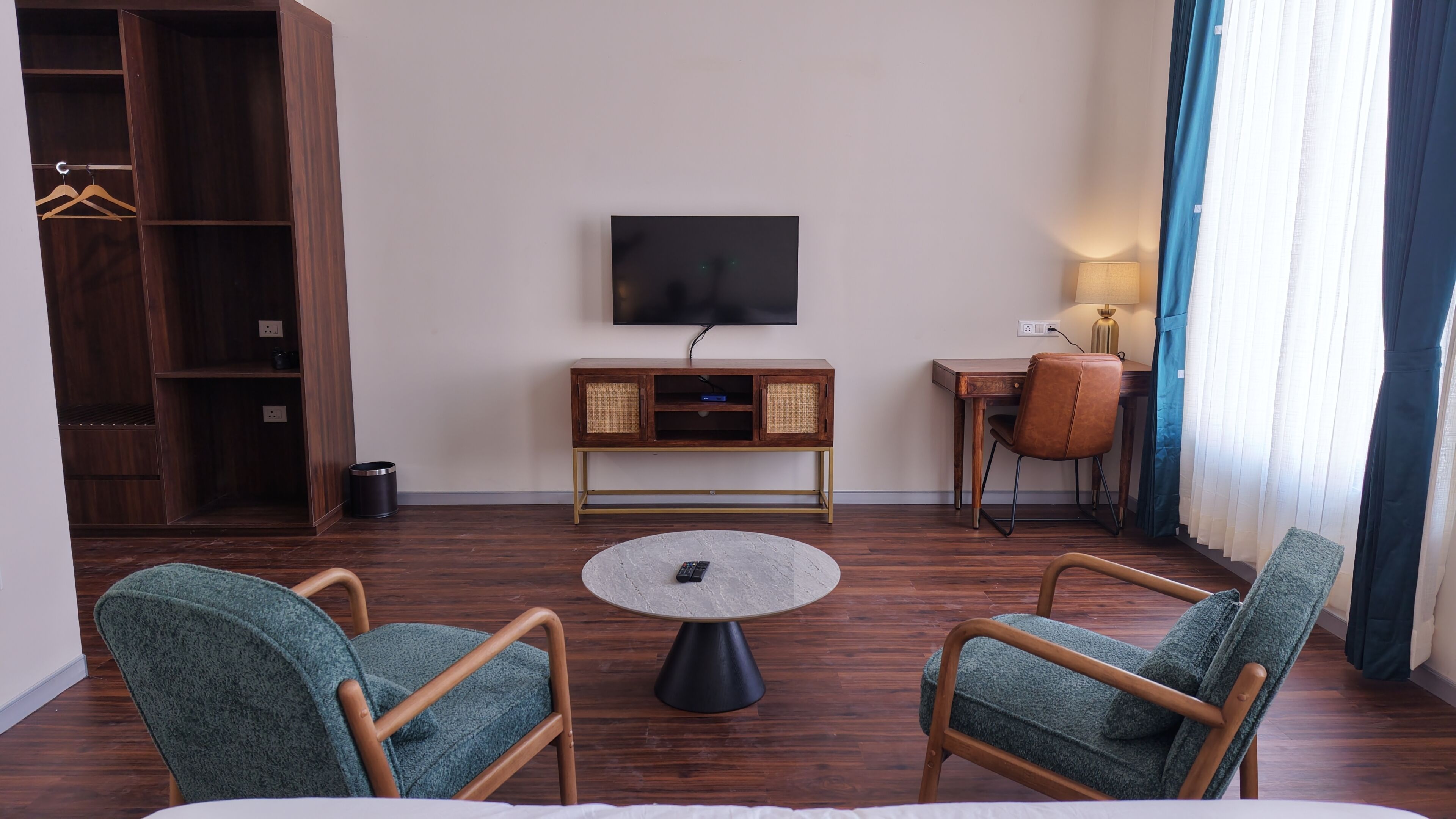 Quarto de hotel com televisão, pequena escrivaninha, cadeira, mesa redonda e guarda-roupa.