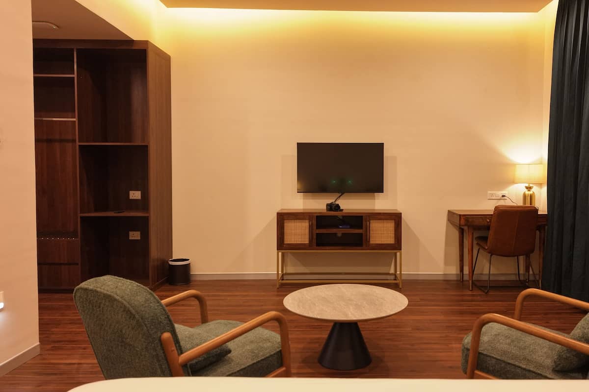 Ein Hotelzimmer mit einem Bett, zwei Sesseln, einem kleinen runden Tisch, einem Fernseher an der Wand, einem Schreibtisch mit einer Lampe und einem Bücherregal.