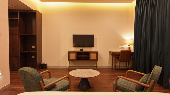 A hotel room with a bed, two armchairs, a small round table, a TV mounted on the wall, a desk with a chair, and a bookshelf.
