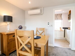 Habitació amb escriptori de fusta, cadira i làmpada. Es veu una cuina a través d'una obertura.