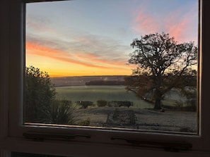 Blick aus dem Fenster auf einen Sonnenuntergang über einer ländlichen Landschaft mit einem großen Baum und offenen Feldern.