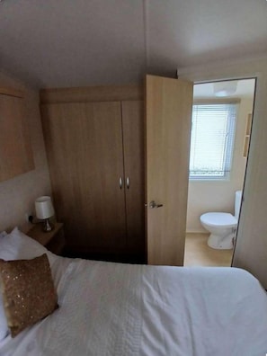A bedroom with a bed, a wardrobe, a nightstand with a lamp, and a view of a bathroom. - Hoburne Cotswolds Retreat (South Cerney)