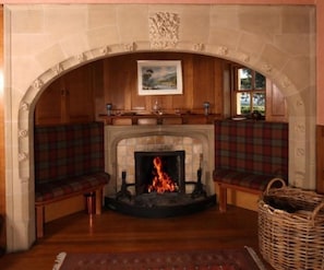 A fireplace with a fire, flanked by two plaid benches, under a stone archway with a painting above. - Glenfarg House, Glenfarg (Glenfarg)