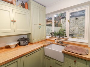 A kitchen with green cabinets, a wooden countertop, a sink, and a window with a view of a brick wall and plants. - The Cottage Loaf, Wedmore (Wedmore)