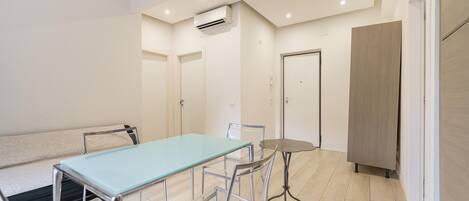 A modern room with a glass-top table, metal chairs, a small round table, a sofa, a wardrobe, and an air conditioning unit.